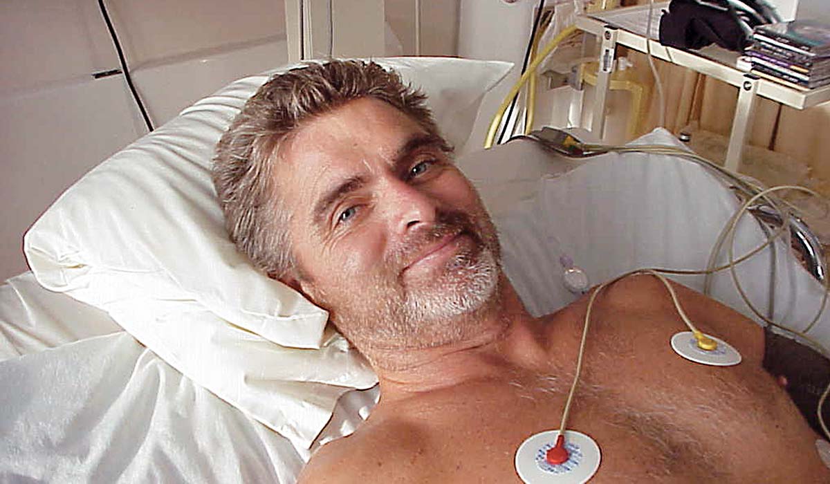 Man lies in hospital bed. He's shirtless and hooked up to electrode pads.