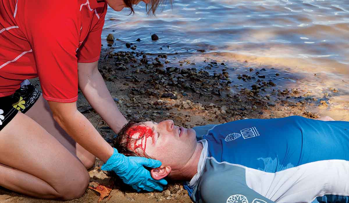 Man with bloodied forehead lies on the beach while a female medic treats him