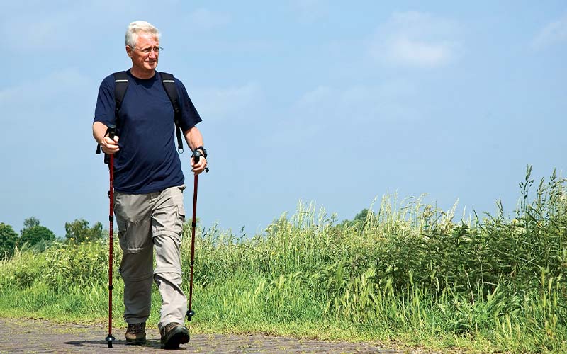 Old man, holding walking sticks, walks a trail in a meadow