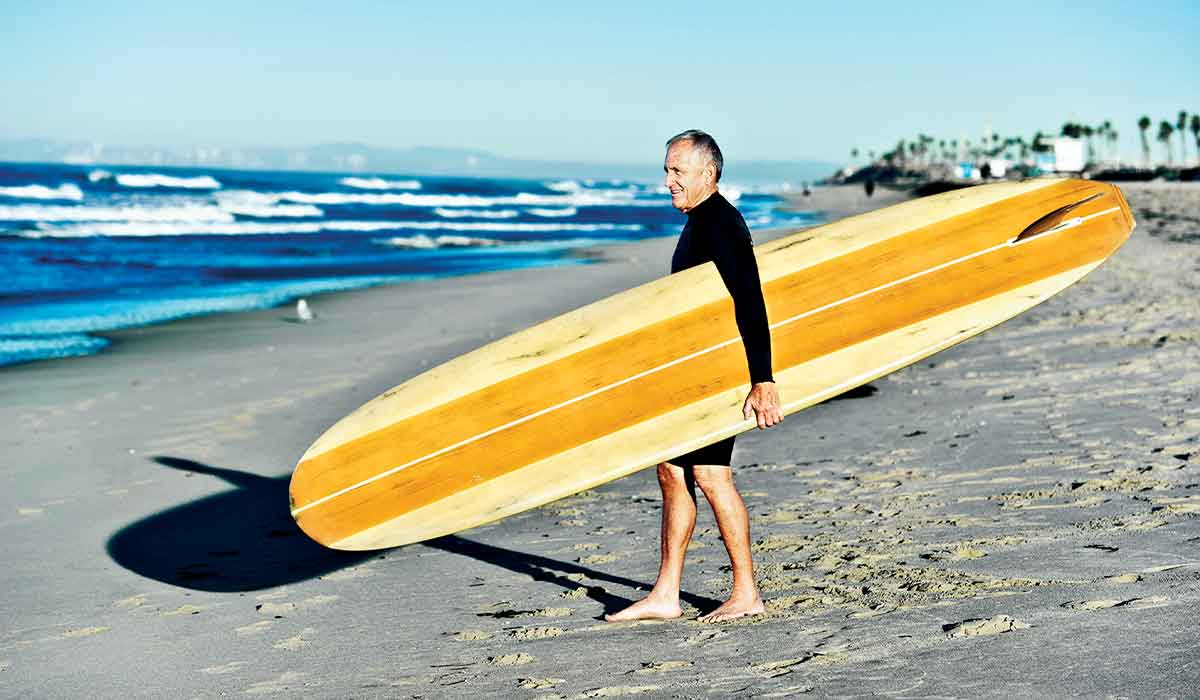 Old man walks across beach holding a yellow surfboard