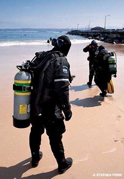 Three drysuit divers walk on the beach toward the ocean to start their dive
