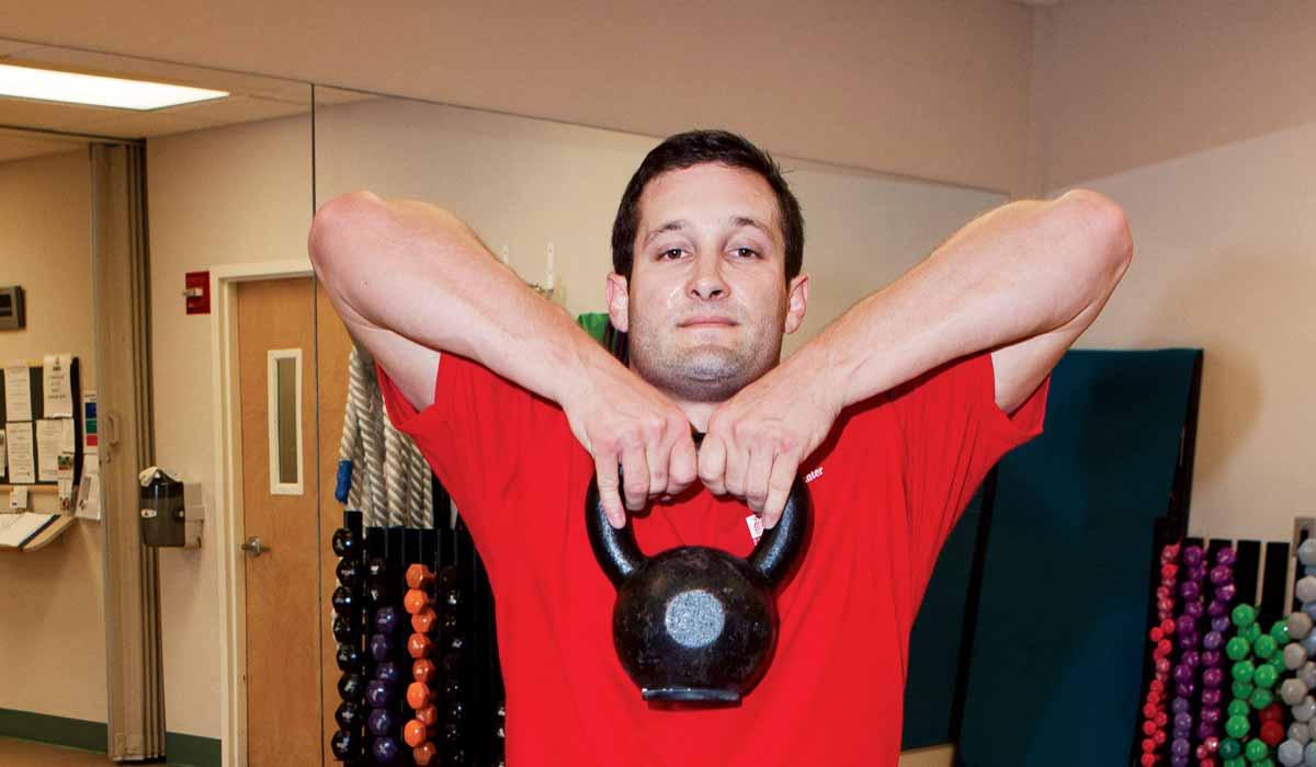 Torso of man in red short holding a kettlebell up to his chin