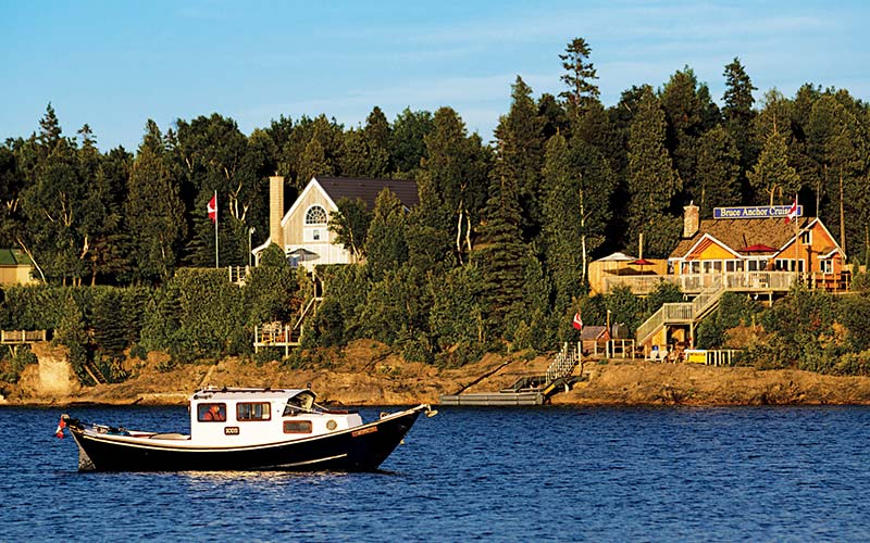 Tugboat floats on the water. A pretty house is in the background on the shore. There are fir trees everywhere. 