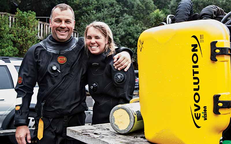A female and male diver stand topside. The man has his arm around the woman and a bright-yellow rebreather tank is in the foreground