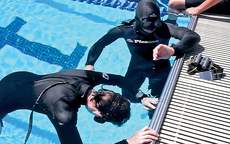 Two freedivers practice in a pool. One is face down in the water holding their breath and the other is timing them.