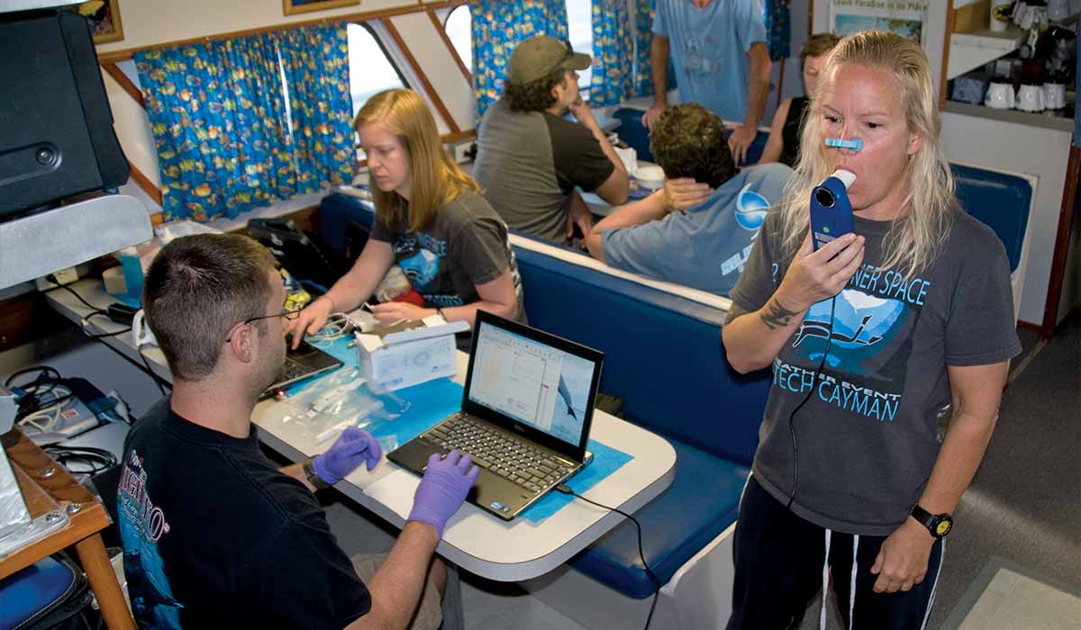 Woman performs respiratory test while man sits nearby tracking progress on laptop