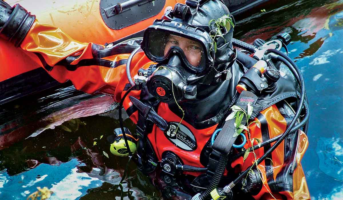 A public safety diver in an orange, weed covered suit has surfaced next to his zodiac.
