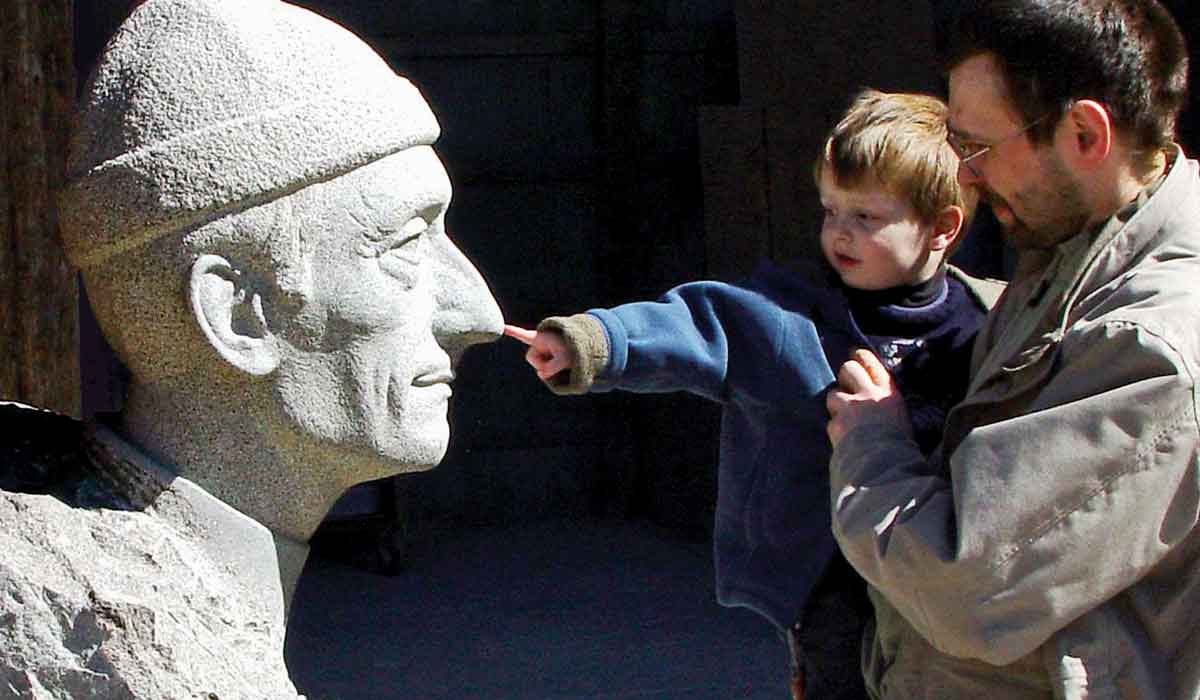 A toddler, held by their father, pokes the nose of a giant stone bust