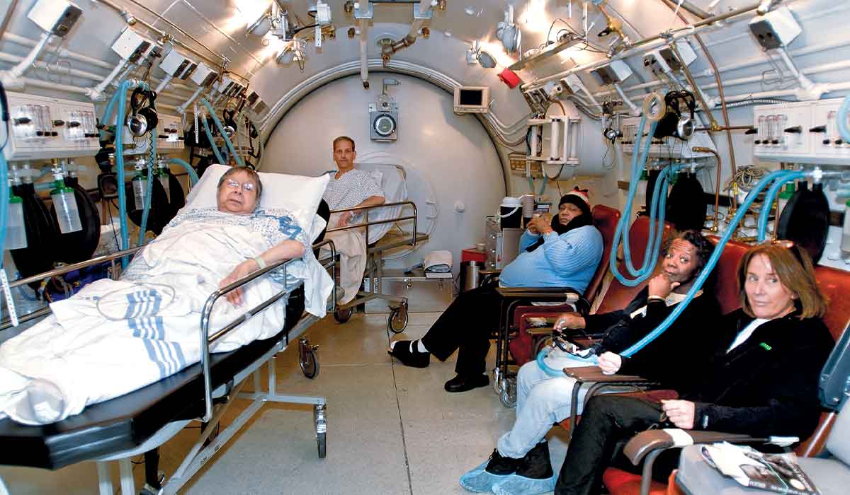 Hyperbaric chamber with five people — three people in chairs and two people in hospital beds.