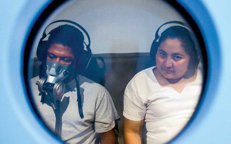 Inside a hyperbaric chamber is a man wearing an oxygen mask and a female aide sits next to him