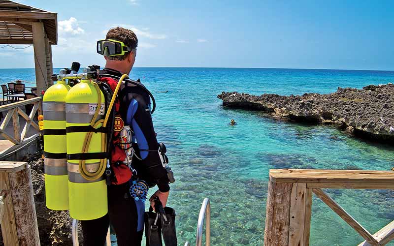Male diver stands on above-ground deck wearing backplate buoyancy system with green oxygen tanks