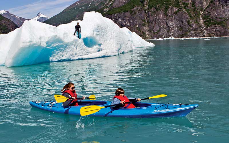 Two people, wearing red life jackets, paddle a blue kayak next to an iceberg