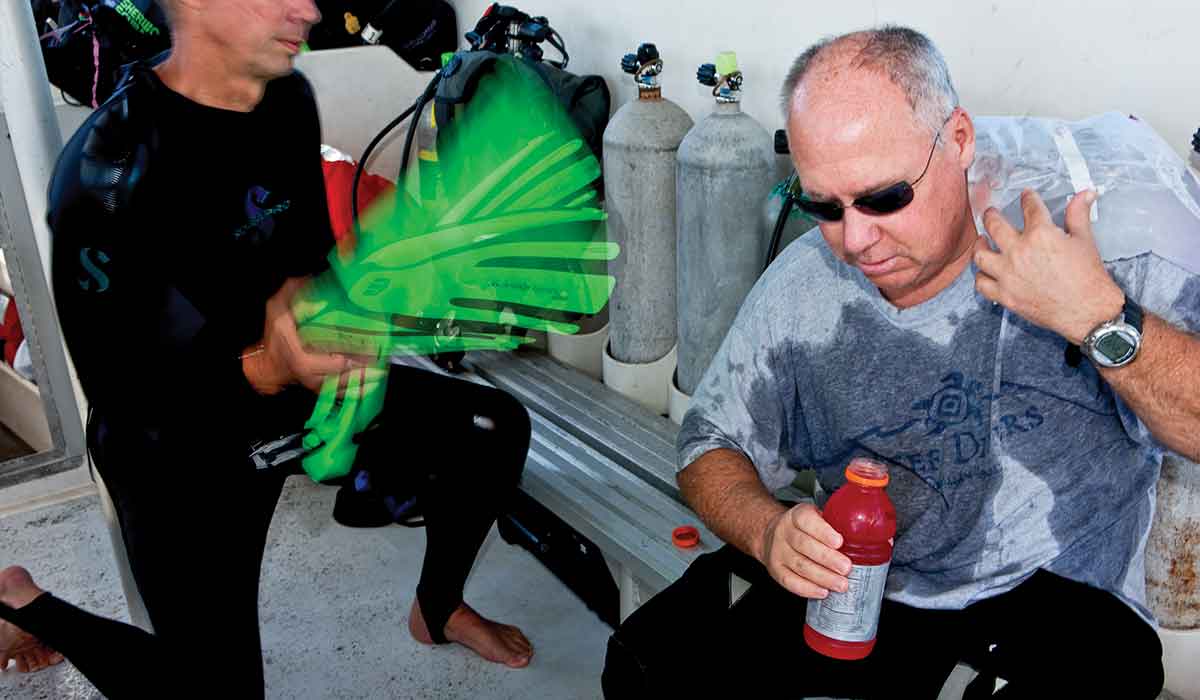 Very sweaty man sits down on boat and is holding a sports drink. A man next to him is kneeling and using lime-green fins to fan the sweaty man