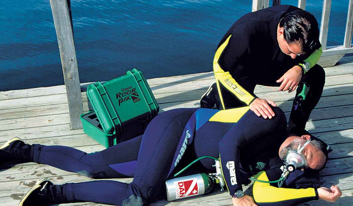 A diver lies on a dock in the recovery position and is wearing an oxygen mask. A second diver is kneeling behind and checking to make sure he's ok.