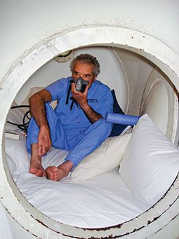 Barefoot man in scrubs sits in a hyperbaric chamber wearing an oxygen mask