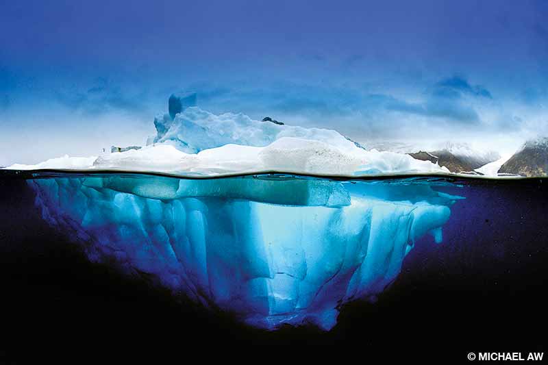 An over-under photo of an iceberg with the fast-eroding mountain glacier in the background at Scoresby Sund, Greenland