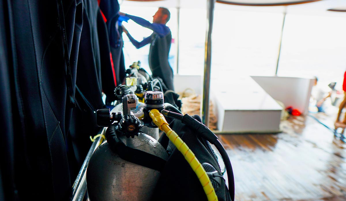 Stored tanks are standing neatly on a dive boat. In the background is a diver preparing some gear