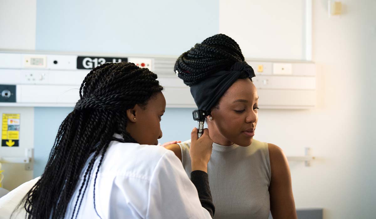 A female Black ear doctor looks into the right ear of a female Black patient.