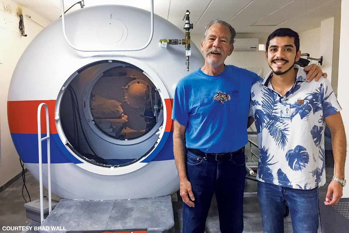 two men smile in front of a hyperbaric chamber