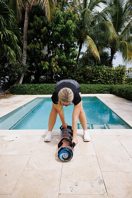 Woman stands with feet hip-width apart and the bag between her legs, with the back of the bag even with her heels. She uses her right hand to grab the bag’s rear handle, which is between her feet.