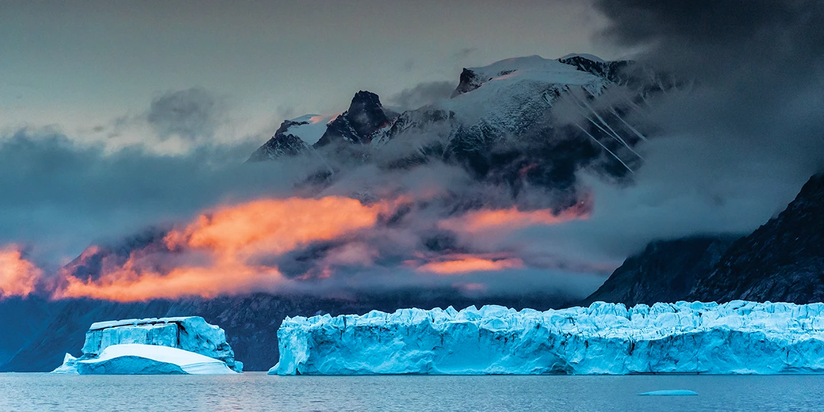Towering peaks trap late autumn light in Scoresby Sund, illuminating a building weather system moving through the jagged peaks of East Greenland.